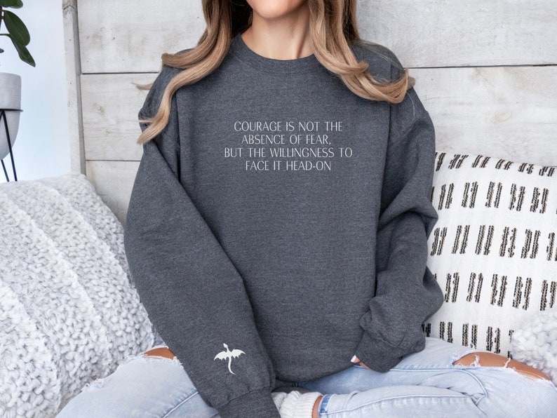May include: A dark gray sweatshirt with a white quote that reads "Courage is not the absence of fear, but the willingness to face it head-on". A white dragon is embroidered on the left sleeve.
