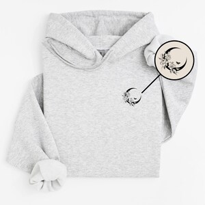 K&ouml;nnte beinhalten: Ein hellgrauer Kapuzenpullover mit einem floralen Halbmond-Design. Das schwarz-wei&szlig;e Design zeigt Blumen und einen Schmetterling. Der Kapuzenpullover hat eine Kapuze und lange &Auml;rmel.