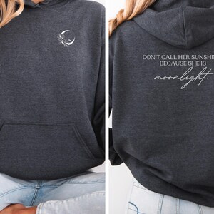 K&ouml;nnte beinhalten: Dunkelgrauer Kapuzenpullover mit Fronttasche und Kapuze. Die Vorderseite zeigt einen wei&szlig;en Halbmond und ein Blumendesign. Auf der R&uuml;ckseite steht der Text "DON'T CALL HER SUNSHINE... BECAUSE SHE IS moonlight" in wei&szlig;er Schrift.