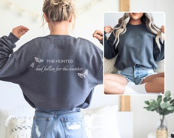 May include: A dark gray sweatshirt with the text "THE HUNTED had fallen for the hunter." printed on the back. The sweatshirt has a relaxed fit and is shown being worn by a person.