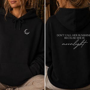 K&ouml;nnte beinhalten: Schwarzer Kapuzenpullover mit Kordelzug und Vordertasche. Auf der Vorderseite befindet sich ein kleiner Mond und ein Blumendesign. Auf der R&uuml;ckseite des Hoodies steht der Text "DON'T CALL HER SUNSHINE. BECAUSE SHE IS moonlight."