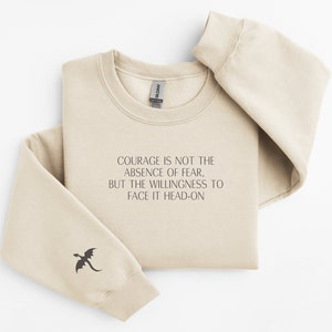 May include: A beige sweatshirt with a black dragon graphic on the left sleeve. The text "COURAGE IS NOT THE ABSENCE OF FEAR, BUT THE WILLINGNESS TO FACE IT HEAD-ON" is printed on the front.
