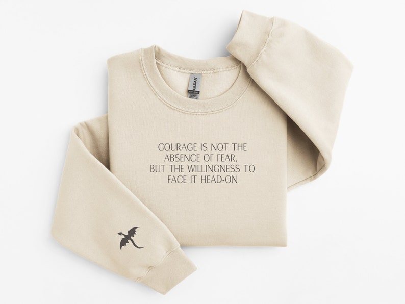 May include: A beige sweatshirt with a black dragon graphic on the left sleeve. The text "COURAGE IS NOT THE ABSENCE OF FEAR, BUT THE WILLINGNESS TO FACE IT HEAD-ON" is printed on the front.