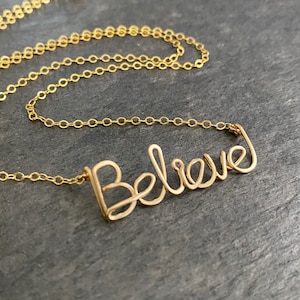 Könnte beinhalten: Eine goldene Kettenkette mit einem zarten Draht-Wortanhänger, auf dem "Believe" steht.