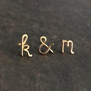 Puede incluir: Tres letras de alambre dorado, "k", "&" y "m", se muestran sobre un fondo oscuro y texturizado. Las letras están en cursiva, con el ampersand centrado entre las otras dos letras. La imagen está bien iluminada, resaltando el brillo del oro.
