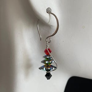 May include: A single silver earring with a Christmas tree design. The tree is made of iridescent, black, red, and green crystals. The earring is hanging from a silver hook.