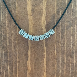 May include: A black cord necklace with silver metal letter beads spelling out "HIJKLMN".
