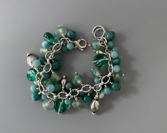 May include: A silver charm bracelet with a variety of green and blue beads. The bracelet features a lobster clasp and a mix of round, oval, and irregular-shaped beads in shades of teal, seafoam, and emerald green.