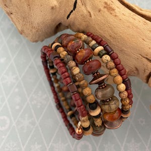 May include: A multi-strand wrap bracelet featuring a variety of beads in shades of deep red, earthy brown, tan, and muted green. The design incorporates smooth round stones, small seed beads, and metallic copper-toned spacers arranged in a spiral.