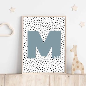 Op de afbeelding: Een blauwe letter M op een witte achtergrond met zwarte stippen. De naam Max staat onder de letter.