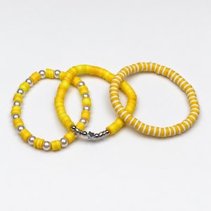 May include: Three bracelets made of yellow clay disc beads. One bracelet features alternating yellow discs and silver-toned beads with a small star charm. Another bracelet alternates yellow discs and white pearls. The third has yellow and white striped discs.