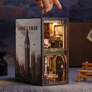 Könnte beinhalten: Miniatur-Diorama aus Holz, das eine Londoner Straßenszene mit einem Uhrturm und der Aufschrift "Rose Road" zeigt. Im Inneren befindet sich ein detailliertes Detektivbüro mit Möbeln und einem Schild mit der Aufschrift "The Detective House".