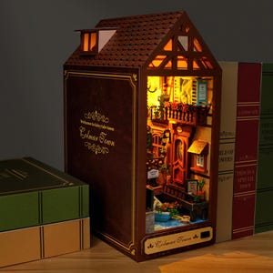May include: A miniature, illuminated model of a European town, designed to resemble a book. The brown exterior features the text "Welcome to fairy tale town Colmar Town". The interior showcases a detailed street scene with miniature buildings and plants.