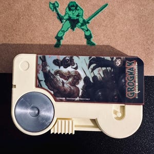 May include: A beige plastic cassette-shaped object featuring a graphic label with the text GROGNAK and a warrior illustration. Behind it sits a brown envelope adorned with a green sticker depicting a muscular figure holding an axe and sword.