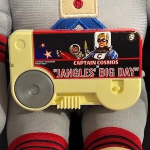 May include: A rectangular, cream-colored plastic game cartridge featuring a graphic label with the text CAPTAIN COSMOS IN JANGLES' BIG DAY. The label depicts a cartoon astronaut and a space character. A circular grey dial is on the left side.