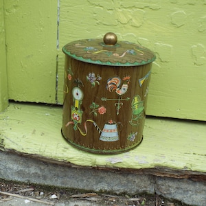 May include: A vintage tin canister with a brown wooden background and colorful illustrations of a rooster, a rocking chair, a watering can, and a clock. The lid is gold and has a knob.