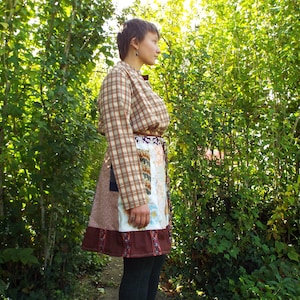 May include: A woman wearing a brown and white plaid shirt with a brown, white, and floral patchwork skirt. She is standing in front of a green bush.