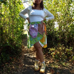 May include: A woman wearing a white top and a cardigan stands in a wooded area. She is wearing a green, yellow, and brown patchwork skirt with a pink and brown fabric pocket. She is wearing gold high-top sneakers.