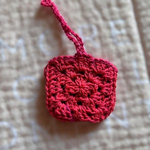 May include: A small, crocheted, pink square with a loop at the top. The square is made of a soft, textured yarn.