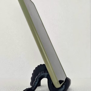 Puede incluir: Un soporte para teléfono negro con forma de pulpo que sostiene un teléfono verde. El soporte está impreso en 3D y presenta un diseño detallado de tentáculos. El teléfono está inclinado para facilitar la visualización.