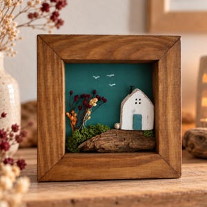 May include: A square wooden shadow box frame containing a miniature scene with a white house, a blue door, green moss, and dried red and yellow flowers against a teal background. Three small white birds are painted in the sky above the house.