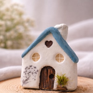 Könnte beinhalten: Ein kleines weißes Keramikhaus mit blauem Dach, herzförmigem Fenster und Holztür. Das Haus hat zwei runde Fenster mit kreuzförmigen Sprossen und einen Schornstein. Auf der Seite ist ein Pusteblumen-Design aufgemalt.