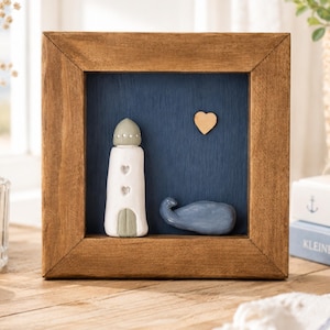 Puede incluir: Una obra de arte enmarcada de temática náutica. Presenta un faro blanco con recortes en forma de corazón, una ballena azul y un corazón de madera sobre un fondo azul oscuro. El marco es de madera marrón, creando una estética rústica.