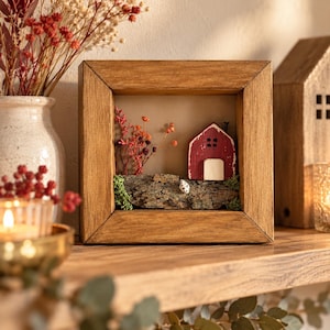 May include: A square wooden shadow box frame containing a miniature red house with a white door, a piece of textured tree bark, green moss, and dried red and orange flowers arranged against a plain light brown background.