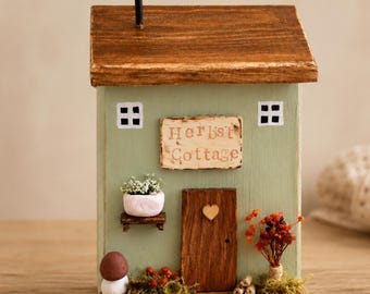 May include: A decorative wooden house figurine painted light green with a dark brown roof and a black nail chimney. It features a central door with a small wooden heart, two white windows, and a sign reading Herbst Cottage. Includes moss and berries.