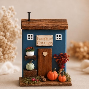 May include: A blue wooden decorative house ornament with a brown roof and a black nail chimney. It features a sign reading Pumpkin Cottage, a central door with a heart accent, two white windows, miniature pumpkins, and dried floral accents.