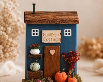 May include: A blue wooden decorative house ornament with a brown roof and a black nail chimney. It features a sign reading Pumpkin Cottage, a central door with a heart accent, two white windows, miniature pumpkins, and dried floral accents.