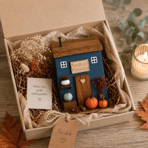 May include: A blue wooden cottage ornament with a brown roof, a sign reading Pumpkin Cottage, and miniature pumpkins. It is packaged in a cardboard gift box with dried flowers, a card with German text, and a brown paper tag labeled F&uuml;r dich.