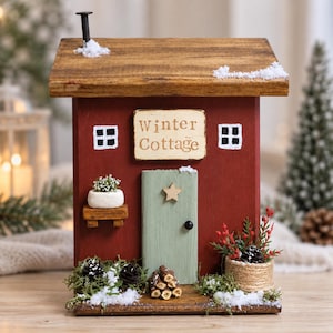 May include: A decorative wooden house model painted red with a brown roof, a light green door, and a sign reading Winter Cottage. It features white painted windows, a small potted plant, a stack of logs, and artificial snow accents.