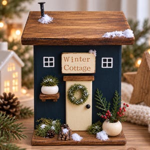 May include: A dark blue wooden cottage decoration with a brown roof, a chimney, and a sign reading Winter Cottage. It features a cream door with a green wreath, two white planters with greenery, a small stack of logs, and artificial snow.