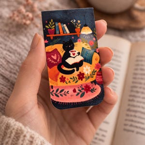 Könnte beinhalten: Rechteckiger Lesezeichen mit einer schwarzen Katze, die ein Buch liest und eine Tasse Kaffee hält. Die Katze sitzt auf einem gelben Sofa mit Kissen, einem Bücherregal und Blumen. Der Hintergrund ist dunkelblau.