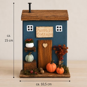 May include: A blue wooden decorative cottage ornament with a brown roof and chimney. It features a sign reading Pumpkin Cottage, a wooden door with a heart, and miniature pumpkins and dried flowers. It measures approximately 15 cm by 10.5 cm.
