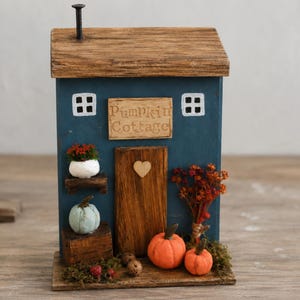 May include: A blue wooden decorative house with a brown roof and a chimney. The front features a sign reading Pumpkin Cottage, a central door with a heart cutout, two white windows, a small potted plant, two orange pumpkins, and dried flowers.