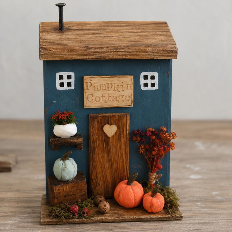 May include: A blue wooden decorative house with a brown roof and a chimney. The front features a sign reading Pumpkin Cottage, a central door with a heart cutout, two white windows, a small potted plant, two orange pumpkins, and dried flowers.