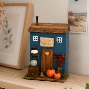 May include: A blue wooden cottage-shaped decoration with a brown roof and a chimney. The front features a sign reading "Pumpkin Cottage," a wooden door with a heart, two white-framed windows, and miniature pumpkins, berries, and moss accents.