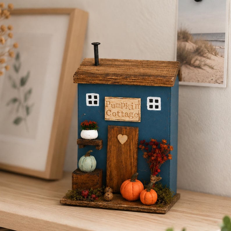 May include: A blue wooden cottage-shaped decoration with a brown roof and a chimney. The front features a sign reading "Pumpkin Cottage," a wooden door with a heart, two white-framed windows, and miniature pumpkins, berries, and moss accents.