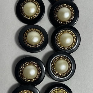 May include: A collection of ten black buttons with a gold rope-like trim surrounding a large, off-white, pearl-like center. The buttons are round and have a glossy finish, arranged in a grid pattern.