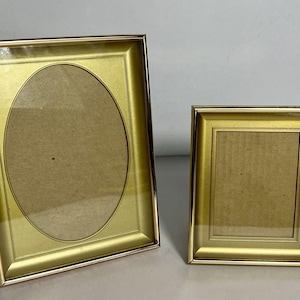 May include: Two gold-toned metal picture frames with a polished finish. The larger frame features a rectangular outer edge with an oval-shaped inner matting, while the smaller frame has a rectangular outer edge with a rectangular inner matting.