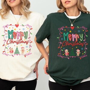 May include: Two t-shirts, one cream and one dark green, each with a festive "Merry Christmas" design. The design includes a heart, lights, snowflakes, and gingerbread figures, perfect for holiday celebrations.
