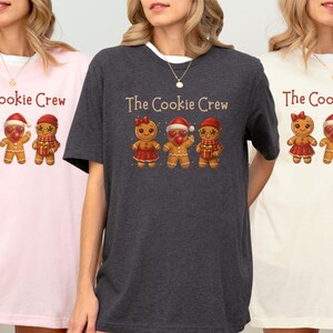 May include: Three t-shirts in pink, charcoal, and cream, each with the text "The Cookie Crew" above a graphic of gingerbread people. The gingerbread people are decorated with bows, Santa hats, and scarves.