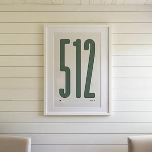 May include: A framed print with the number "512" in a bold, green font. The artwork is centered on a white wall with horizontal paneling. Two beige armchairs are positioned in front of the wall.