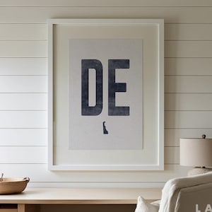 May include: A white framed print with the letters "DE" in black and a silhouette of the state of Delaware in gray. The print is hanging on a white shiplap wall.