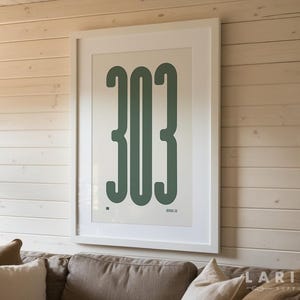 May include: A framed wall art print featuring the number "303" in a bold, dark green font against a white background. The artwork is displayed in a white frame and hangs on a light wood-paneled wall. The bottom of the image shows a section of a sofa with pillows.