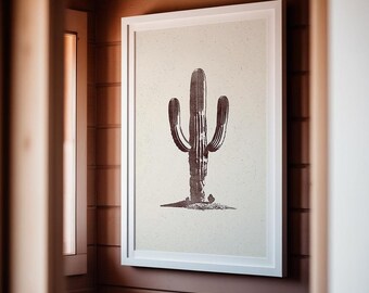 May include: A framed print of a black and white illustration of a cactus on a beige background. The frame is white and the print is hanging on a wooden wall.