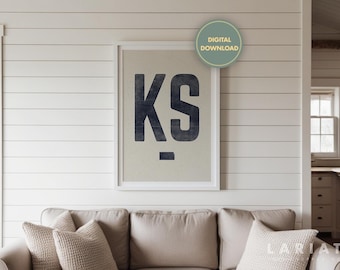 May include: A white framed print with the letters "KS" in black on a beige background. The print is hanging on a white shiplap wall above a beige couch.