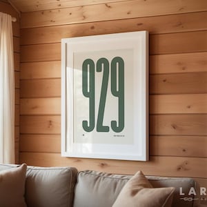 Könnte beinhalten: Ein gerahmter Kunstdruck mit der Nummer "929" in einer kräftigen, grünen Schrift. Der Druck wird an einer Holzwand über einem beigen Sofa präsentiert. Der Raum hat eine gemütliche, natürliche Ästhetik mit einem Fenster und weißen Vorhängen.